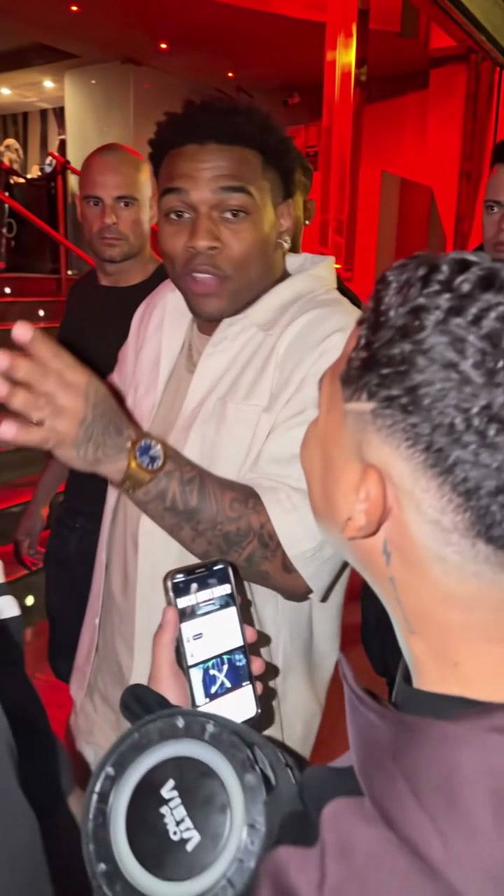 A fictional adult performer in a light shirt gestures in a crowd outside a red-lit entrance