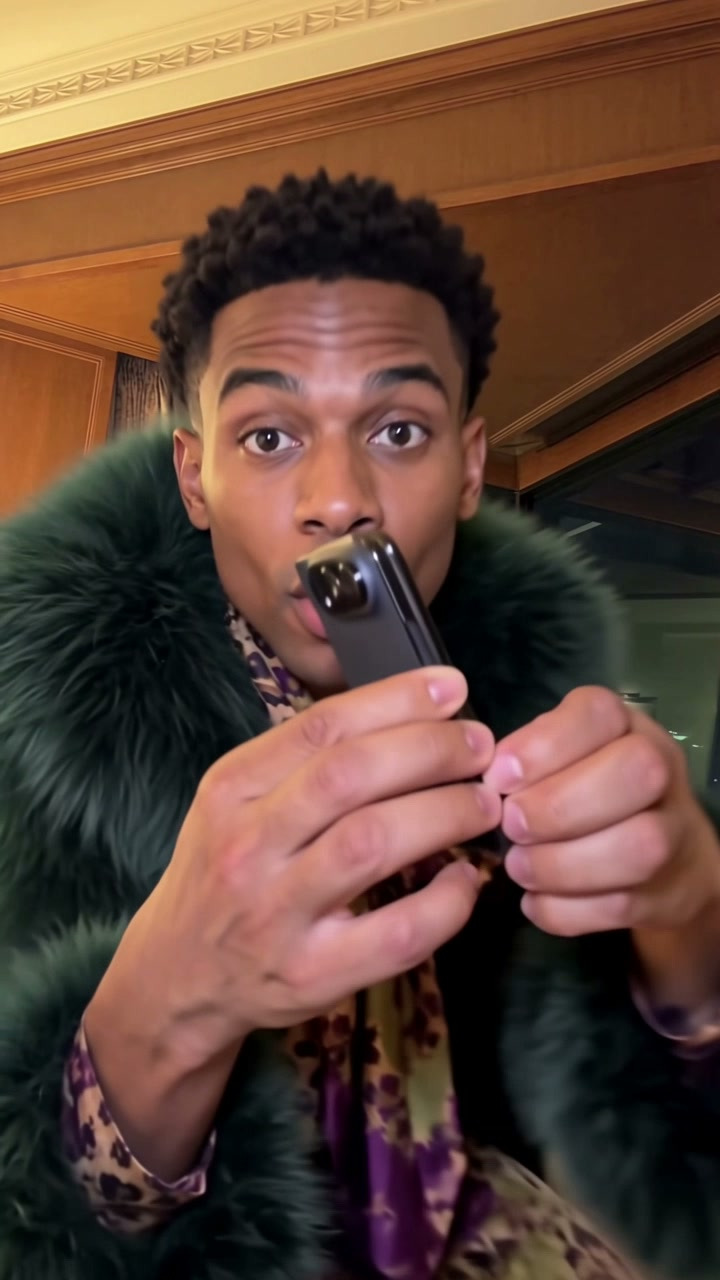 A fictional adult performer in a green coat opens a phone toward the camera in the generated iPhone Kupi preview.